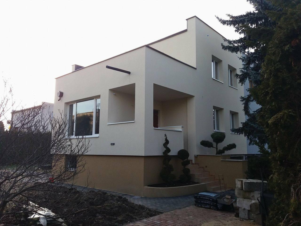 Dwukondygnacyjny dom z jasną elewacją i ciemniejszym cokołem, widoczne schody wejściowe i ozdobne krzewy, na pierwszym planie niezabudowany teren.