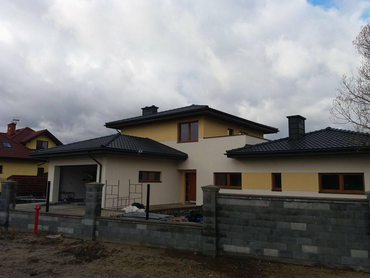 Nowoczesny dom jednorodzinny z elewacją w odcieniach beżu i żółci, grafitowy dach, częściowo ogrodzony murem z bloczków betonowych, widoczne rusztowania i elementy wyposażenia budowy.