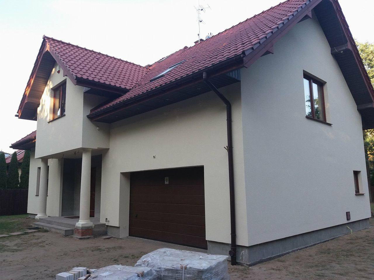 Dwukondygnacyjny dom jednorodzinny z jasną elewacją, ciemnobrązową stolarką okienną i dachem z czerwonej dachówki, widok z poziomu gruntu na fragment elewacji i garaż.