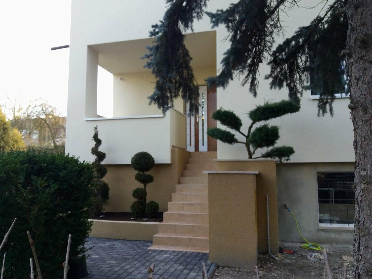 Jasna elewacja domu z wejściem po schodach, ozdobne krzewy, widoczny wąż ogrodowy przy ścianie.