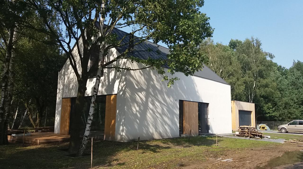 Nowoczesny, biały dom z ciemnym dachem, otoczony drzewami, z widocznym drewnianym tarasem w trakcie budowy i materiałami budowlanymi obok garażu.