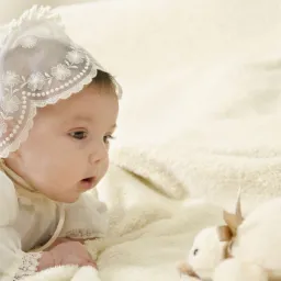 Niemowlę w białym, koronkowym czepku i ubranku, leżące na puszystym kocu, zafascynowane pluszowym misiem z kokardką.