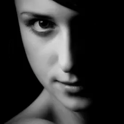 Portret kobiety w czerni i bieli, z połową twarzy skrytą w głębokim cieniu, eksponujący wyraziste oko i delikatny uśmiech.