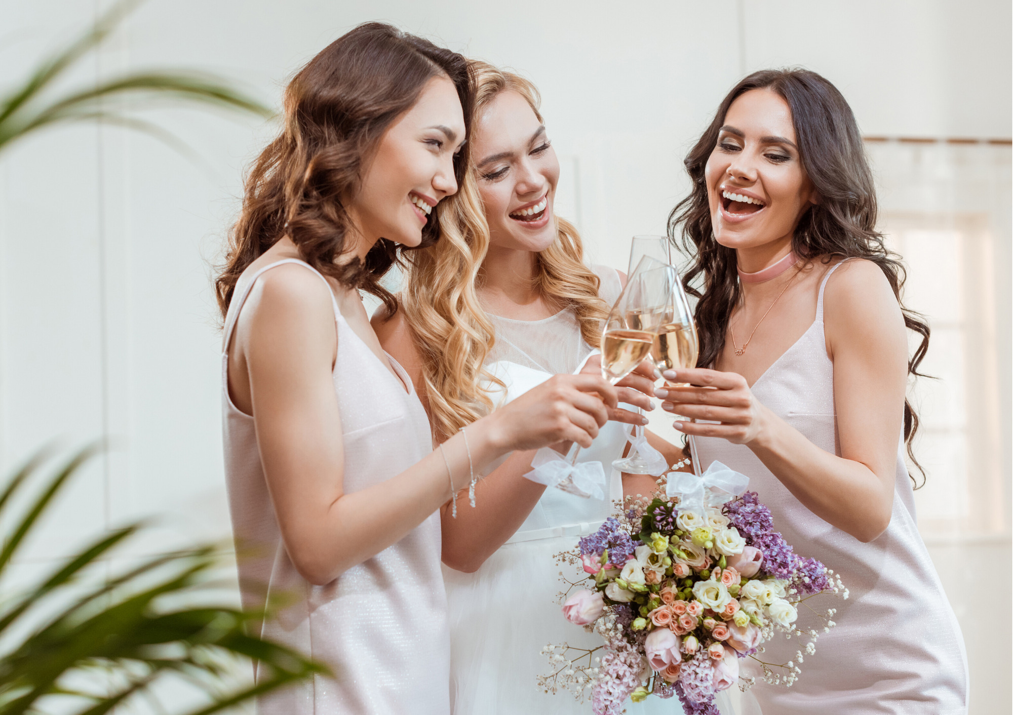 Panna młoda i dwie druhny w jasnych sukienkach wznoszą toast szampanem, trzymając bukiet z różowych i fioletowych kwiatów.