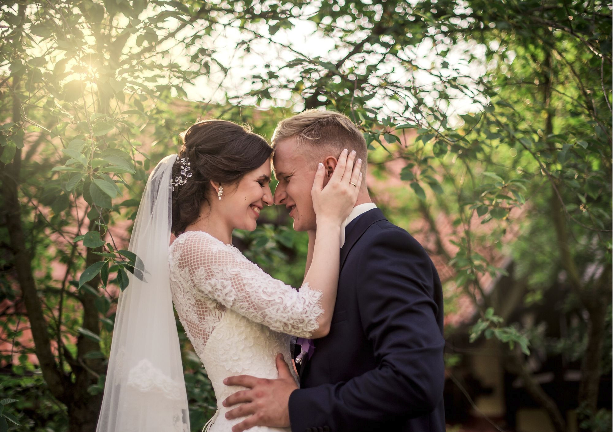 Uśmiechnięta para młoda, panna młoda z welonem i ozdobą we włosach, pan młody w ciemnym garniturze, stykają się czołami na tle zieleni i promieni słonecznych.
