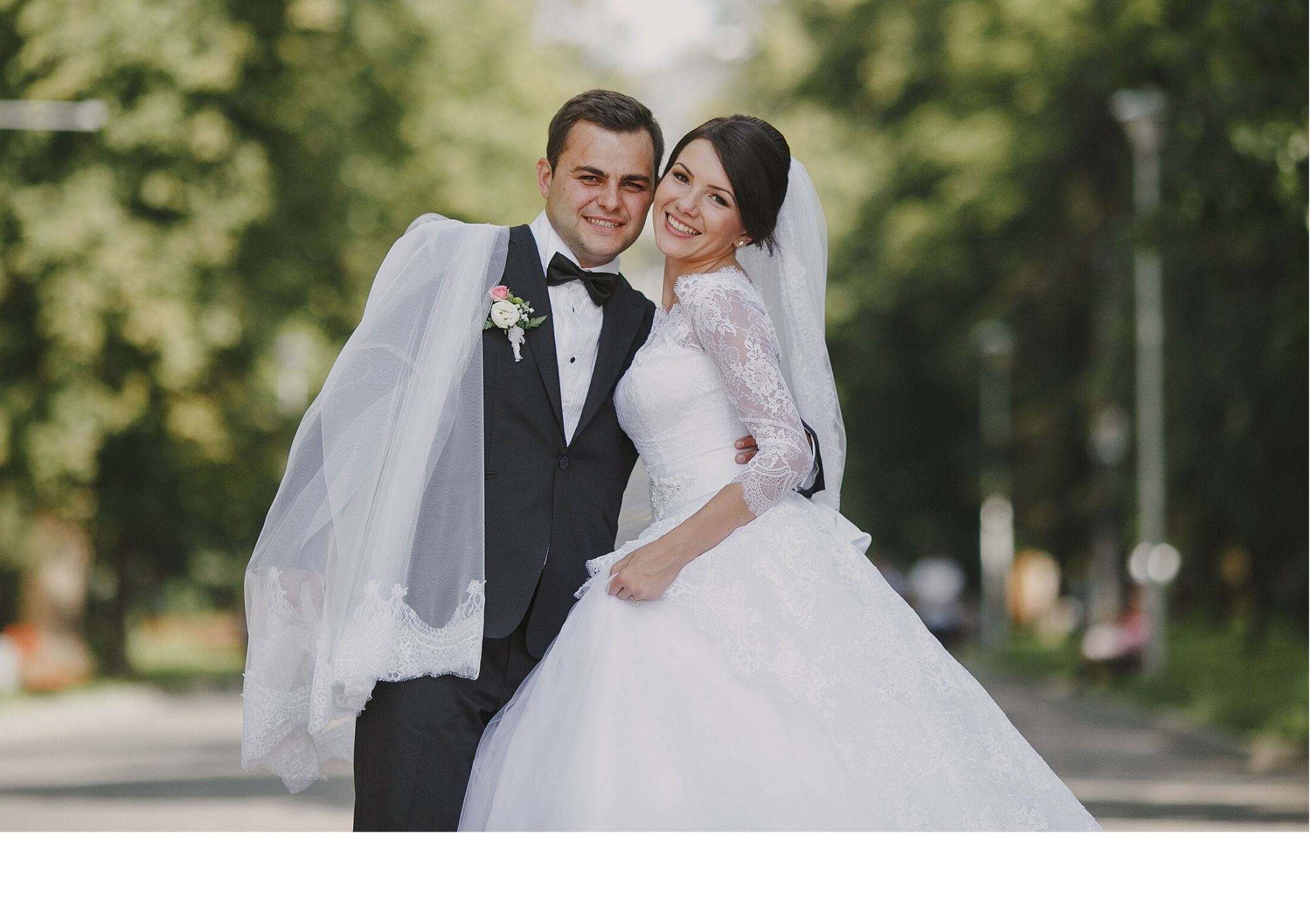 Uśmiechnięta para młoda w plenerze: panna młoda w koronkowej sukni z rękawami 3/4 i welonem, pan młody w czarnym smokingu z muszką i butonierką, welon zarzucony na ramię pana młodego.