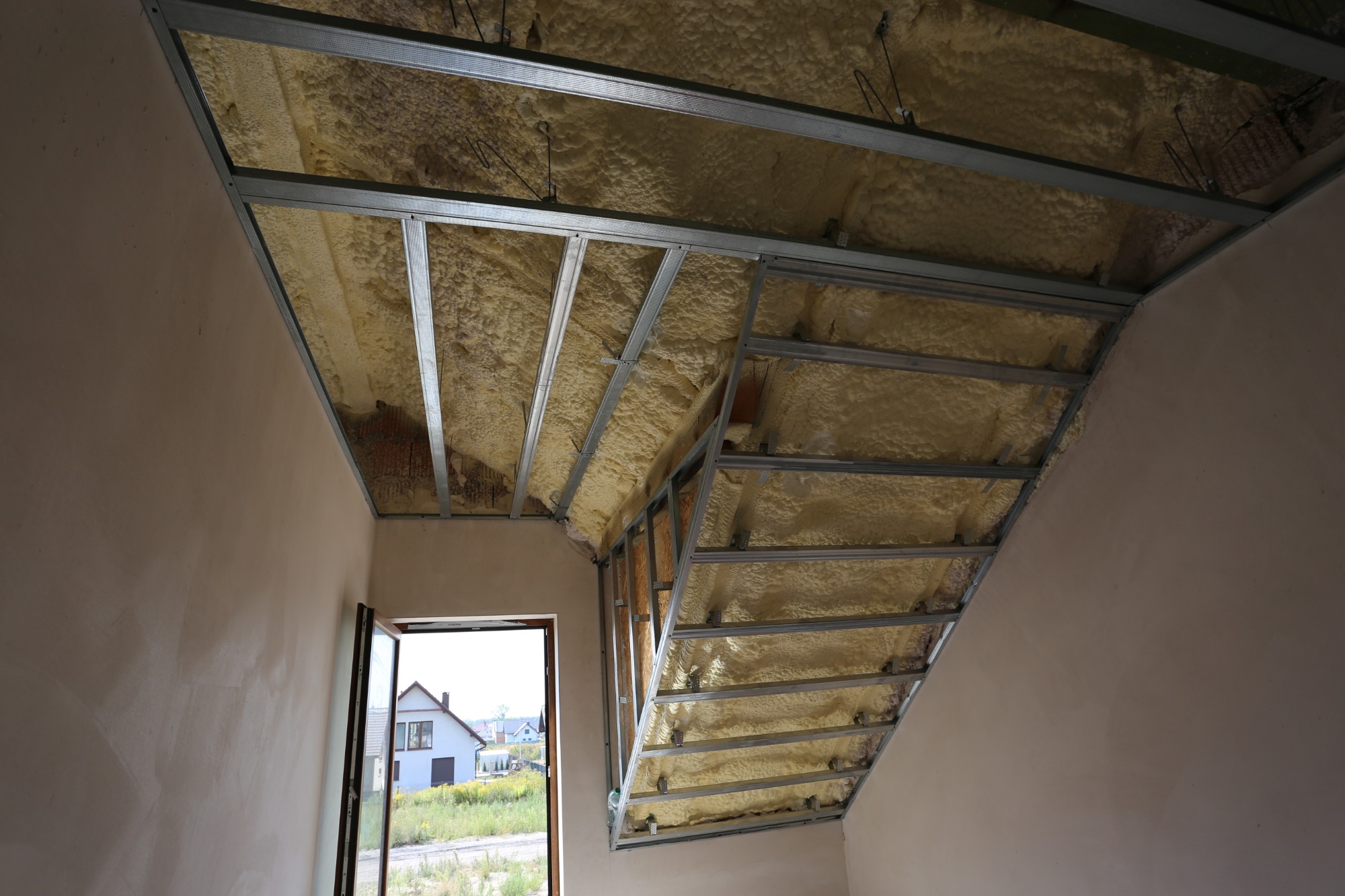 Poddasze z widoczną konstrukcją metalową i nałożoną żółtą pianką izolacyjną, widok z dołu na skośny sufit, otwarte okno z widokiem na dom.