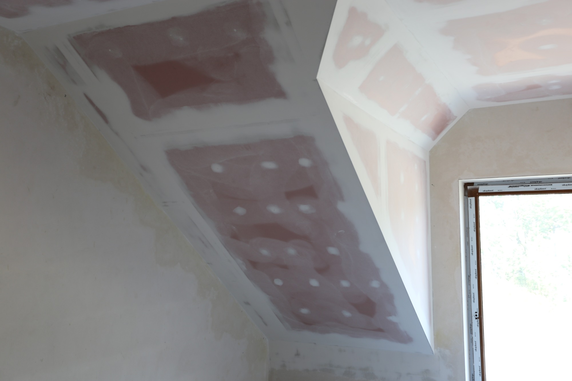 Skośny sufit i ściany w trakcie szpachlowania, widoczne ślady po wkrętach na suficie, okno z widokiem na zieleń w prawym rogu.