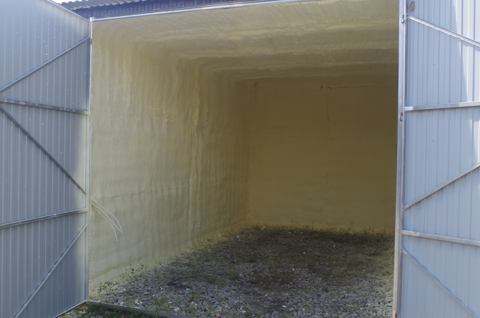 Wnętrze blaszanego garażu po ociepleniu pianką poliuretanową, widoczne otwarte, metalowe wrota i nierówna powierzchnia izolacji na ścianach i suficie, na podłodze żwir.