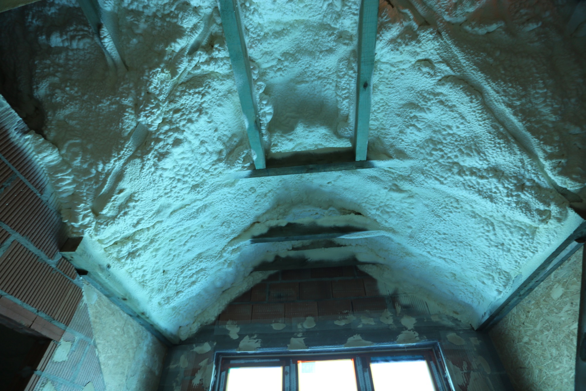 Poddasze z drewnianą konstrukcją pokryte grubą warstwą białej pianki izolacyjnej, widoczne cegły i okno w tle.