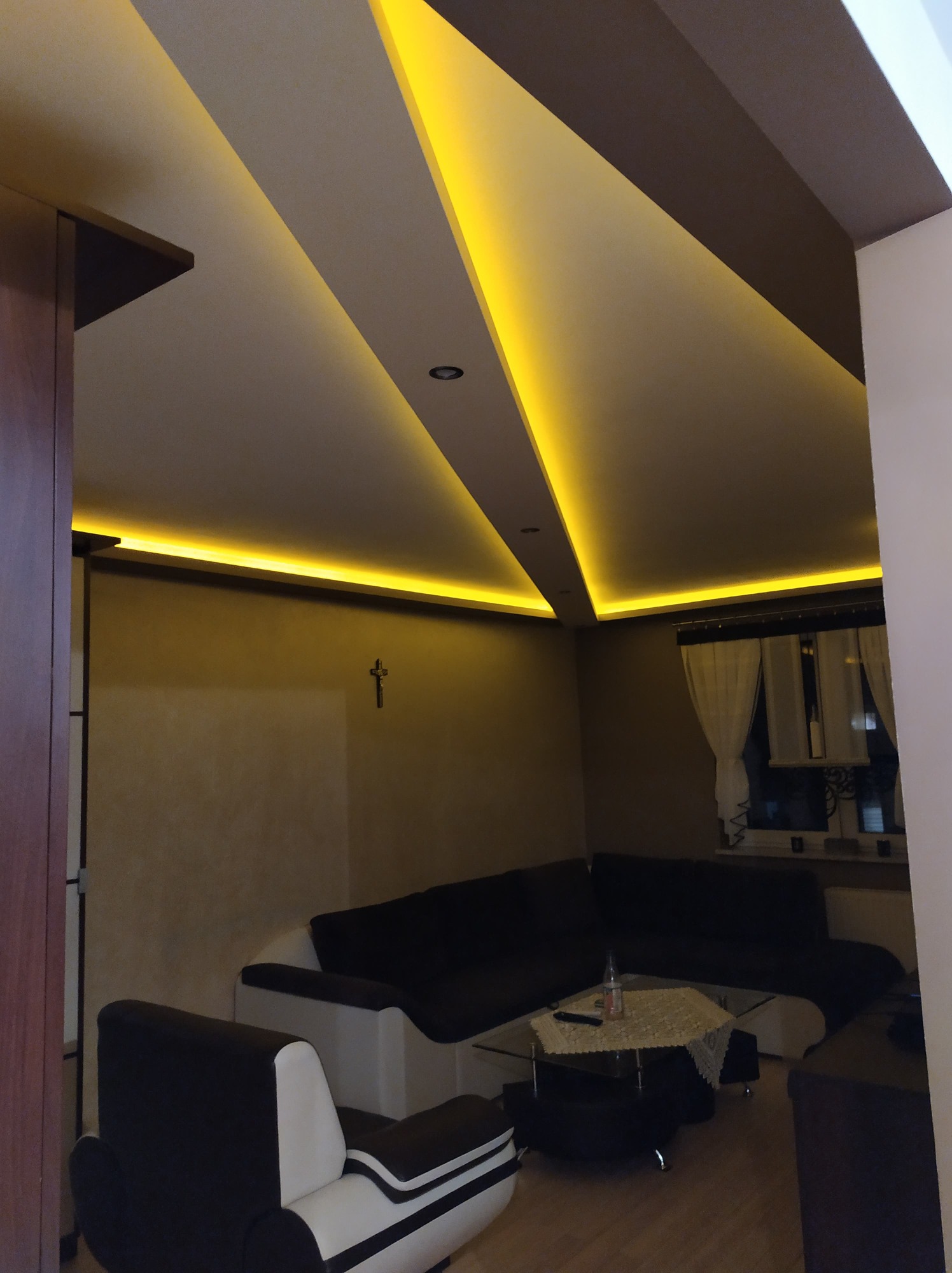 Nowoczesny salon z podświetlanym sufitem wielopoziomowym, oświetlenie LED podkreśla geometrię pomieszczenia, widoczny fragment czarnej sofy i fotela.