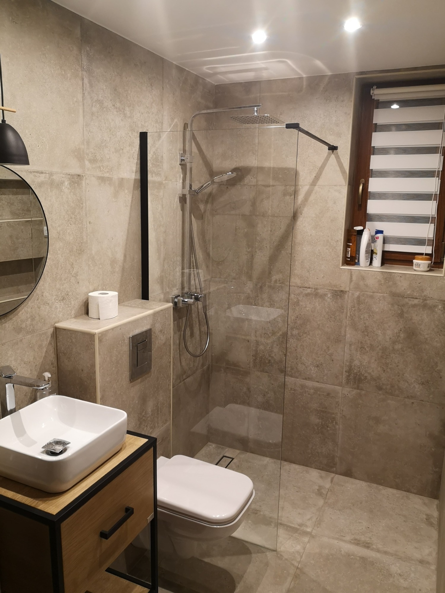 Nowoczesna łazienka z kabiną prysznicową typu walk-in, umywalką nablatową na drewnianej szafce z czarnymi detalami i oknem z żaluzjami.