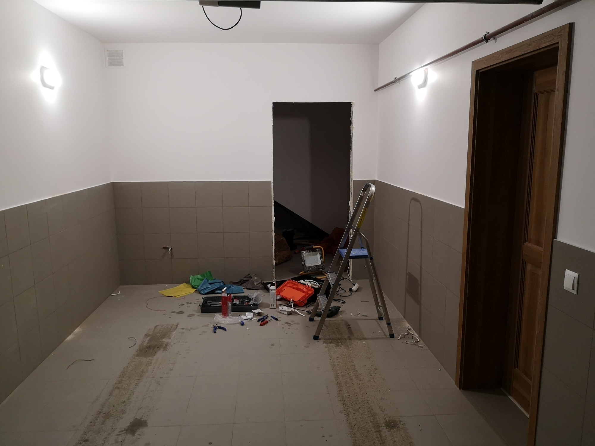 Wnętrze w trakcie prac remontowych: białe ściany z szarym pasem płytek, rozstawiona drabina, narzędzia i akcesoria budowlane rozłożone na podłodze, widoczne ślady po pracy. Otwór drzwiowy bez...