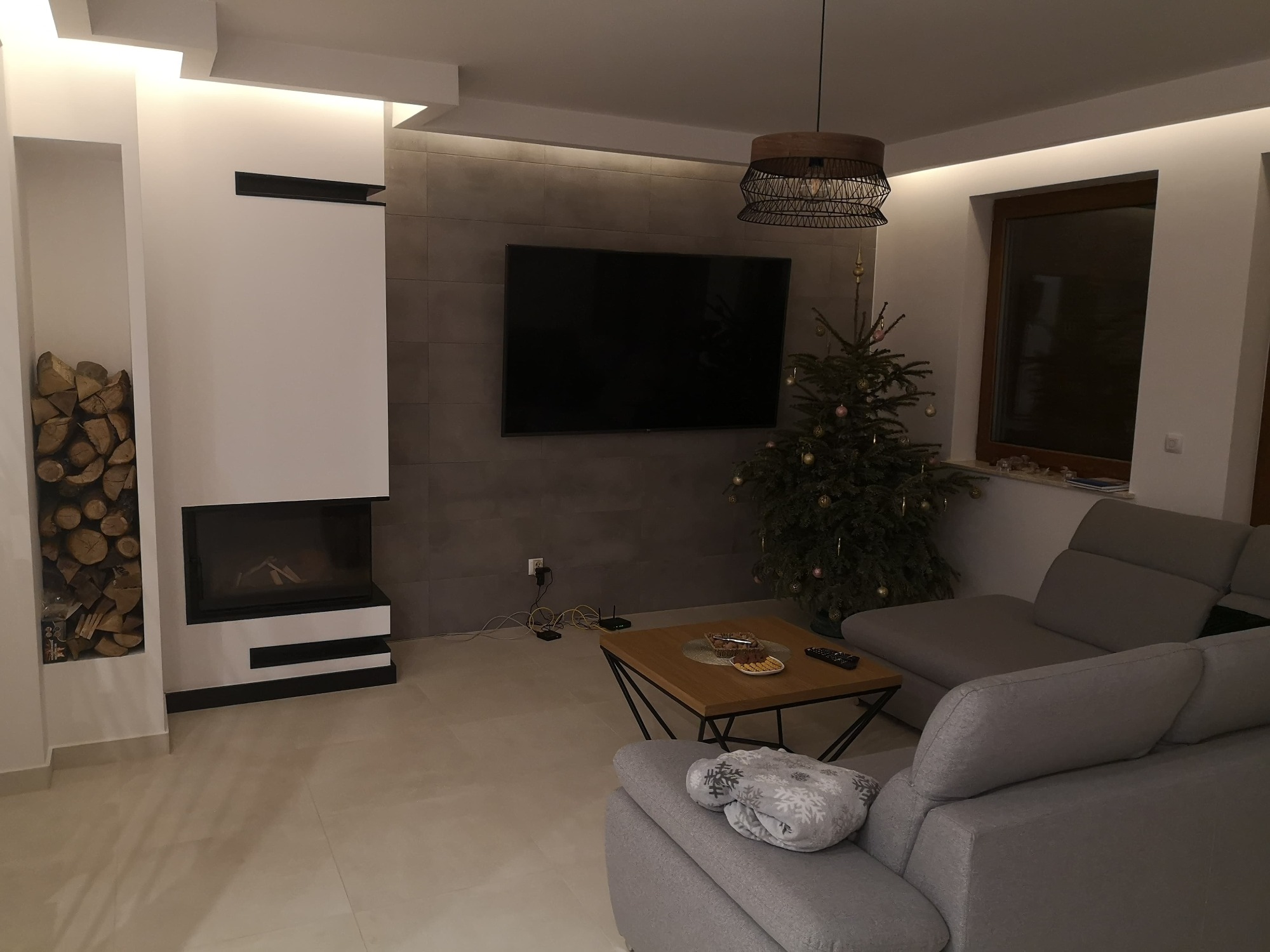Nowoczesny salon z kominkiem, telewizorem na ścianie z szarych płytek i szarą sofą, oświetlony listwami LED pod sufitem, z dekoracjami świątecznymi w postaci choinki.