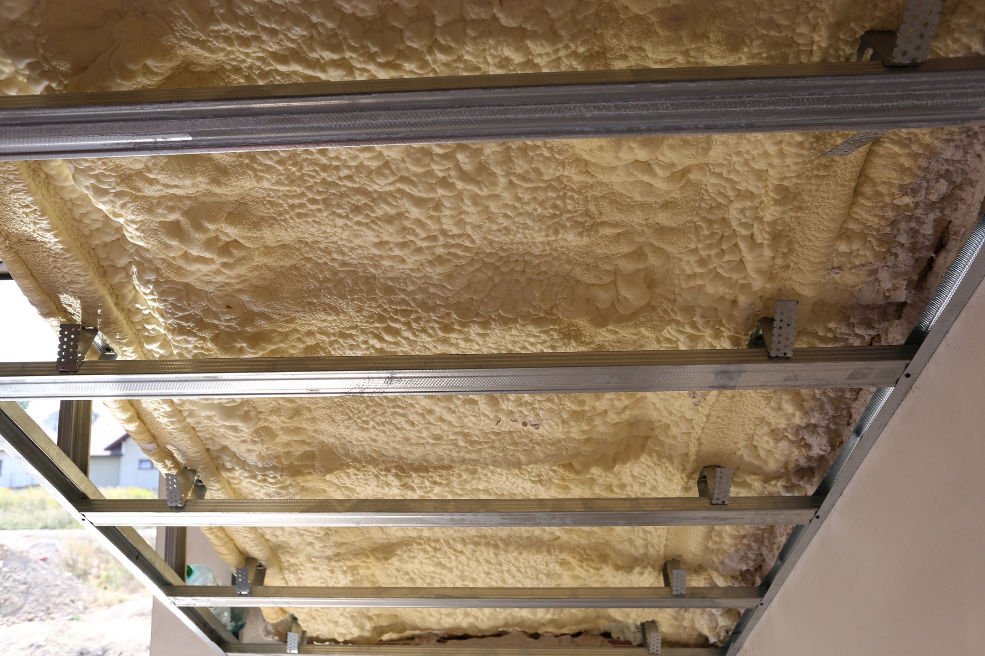 Poddasze ocieplone pianką PUR, widoczne metalowe profile konstrukcyjne, z fragmentem budynku w tle.