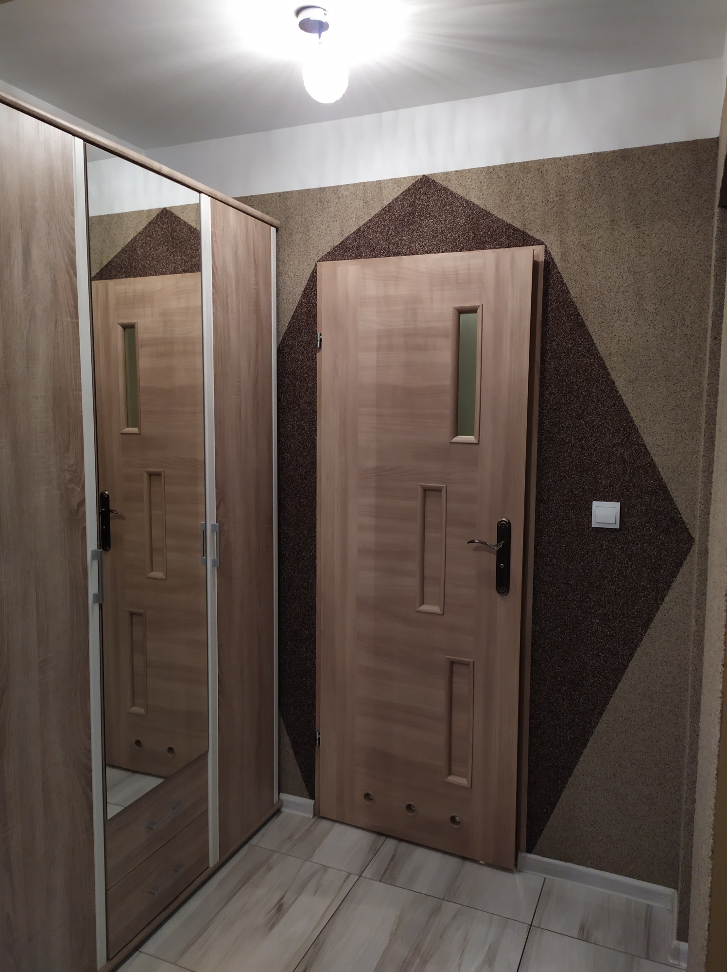 Nowoczesne wnętrze w Zabrzu: drzwi wewnętrzne z przeszkleniem, szafa z lustrem i dekoracyjna ściana z geometrycznym wzorem w odcieniach brązu i beżu.