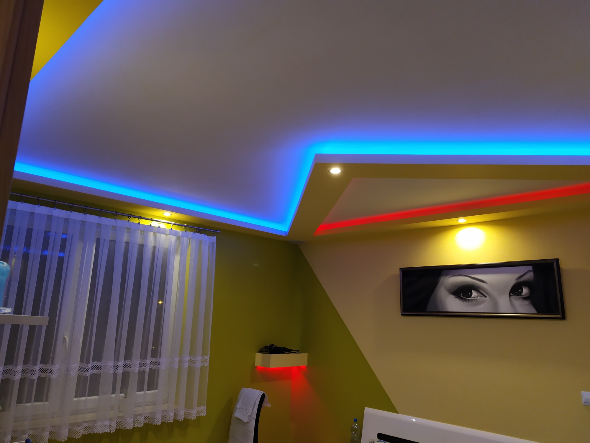 Żółto-biały pokój z podświetlanym na niebiesko i czerwono sufitem, białe firanki przy oknie i obraz z oczami na ścianie.
