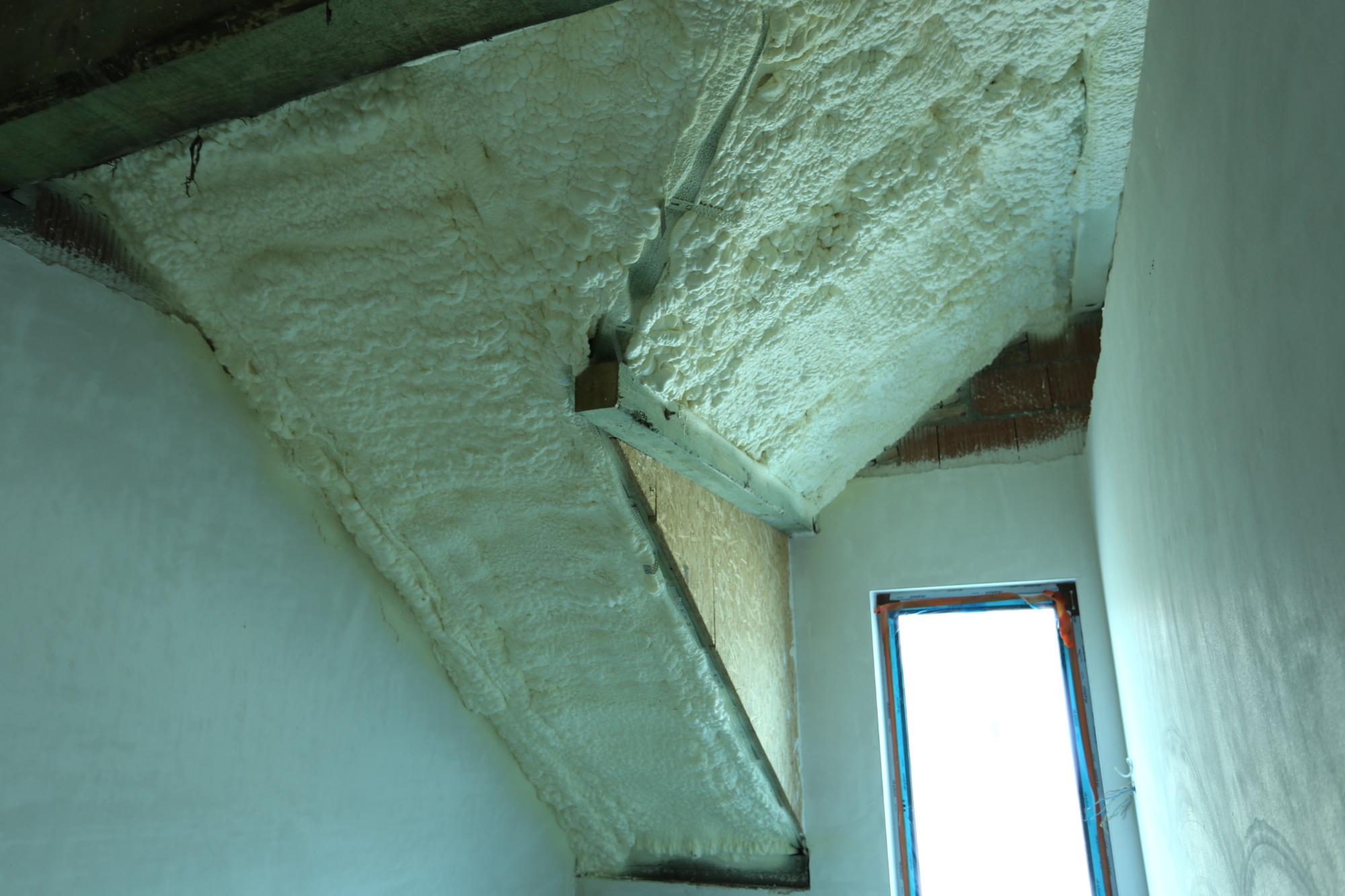 Poddasze z ukośnymi belkami stropowymi pokryte grubą warstwą jasnej pianki izolacyjnej, widoczne fragmenty muru z czerwonej cegły i okno w tle.