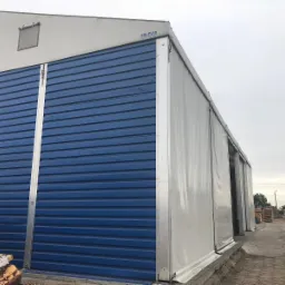 Duży, niebieski, blaszany magazyn z przesuwanymi drzwiami i białym dachem plandekowym, na zewnątrz składowane drewno