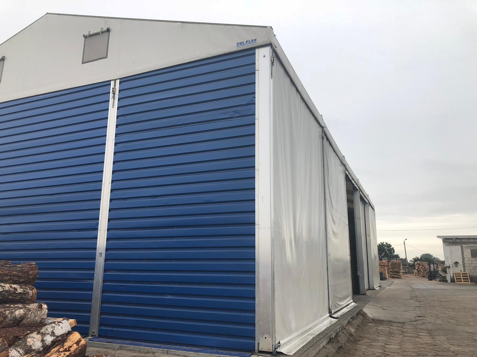 Duży, niebieski, blaszany magazyn z przesuwanymi drzwiami i białym dachem plandekowym, na zewnątrz składowane drewno