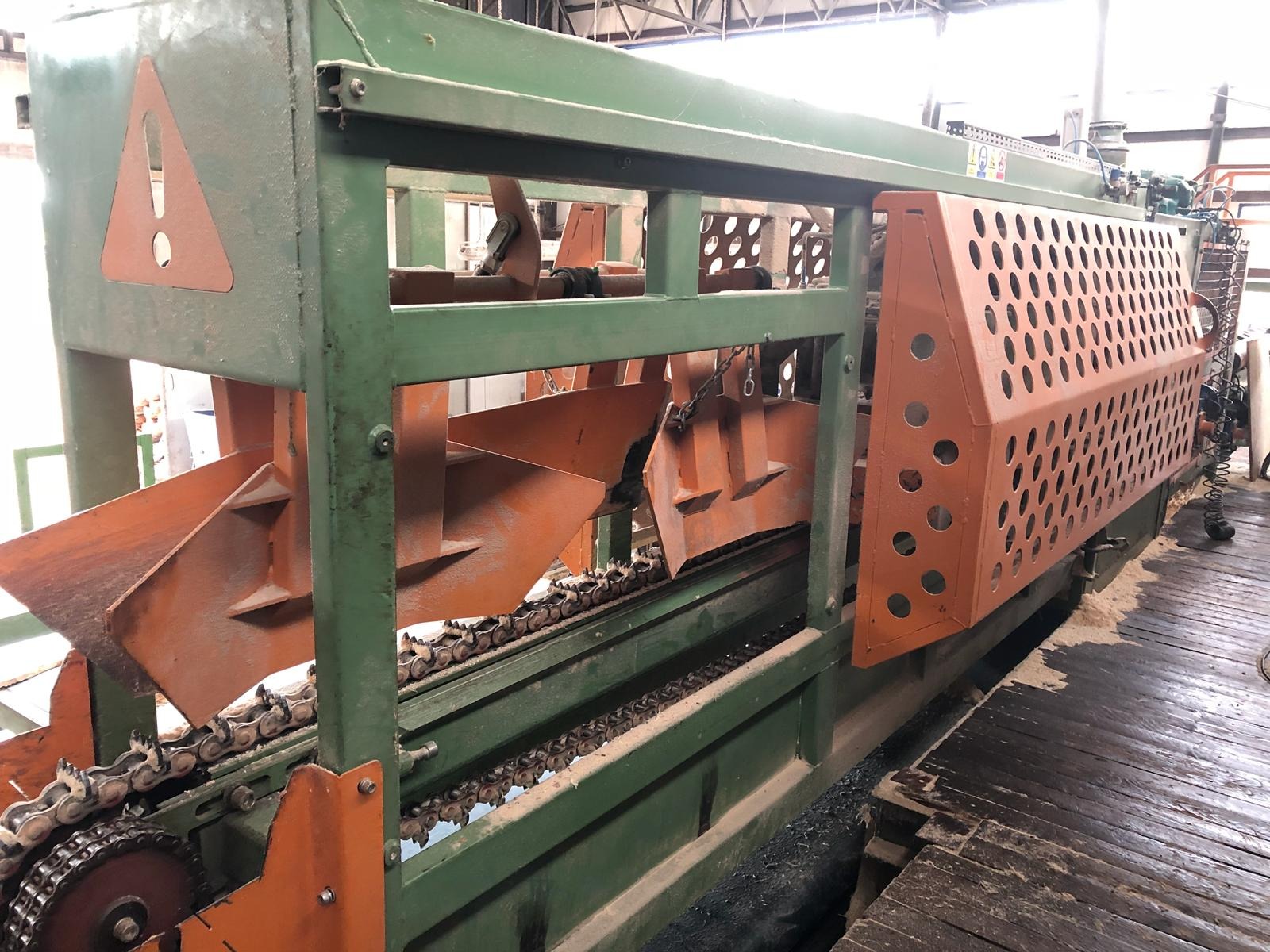 Zielona maszyna do obróbki drewna z pomarańczowymi elementami tnącymi i łańcuchem transportującym, widoczne trociny na podłodze.