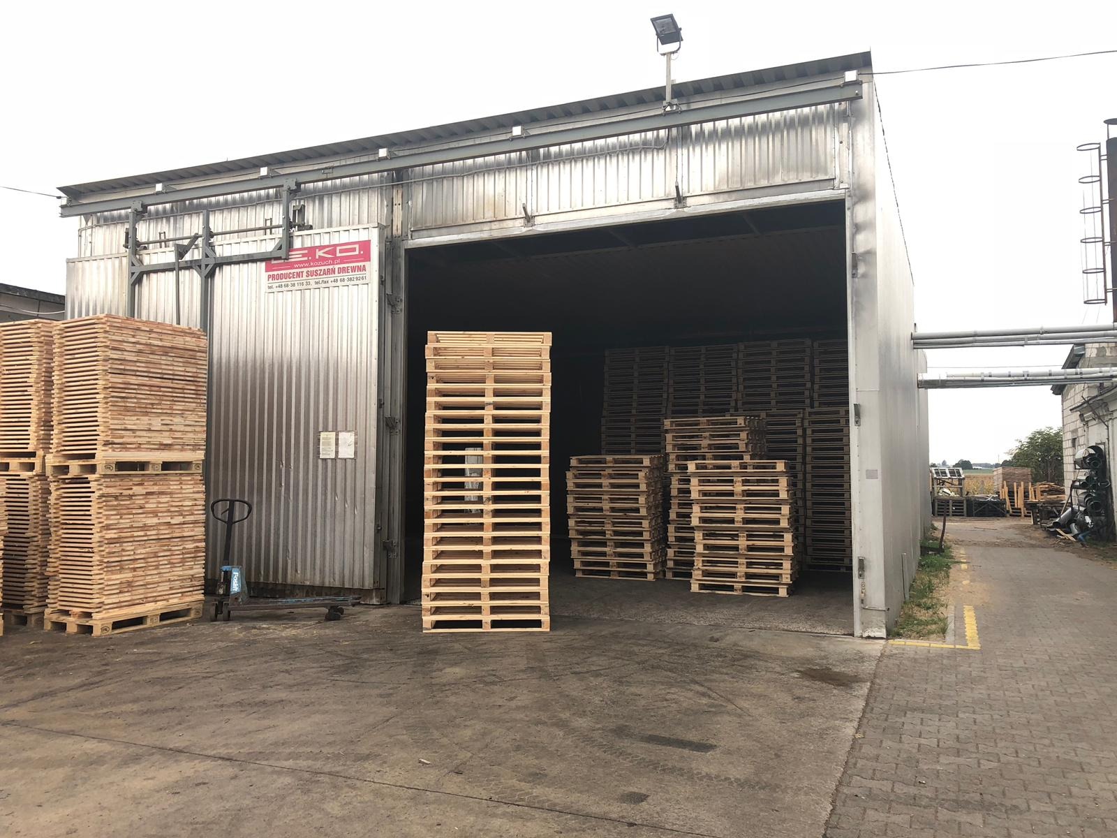 Stosy jasnych, drewnianych palet w magazynie z metalowymi ścianami, widoczny wózek paletowy i logo producenta na ścianie