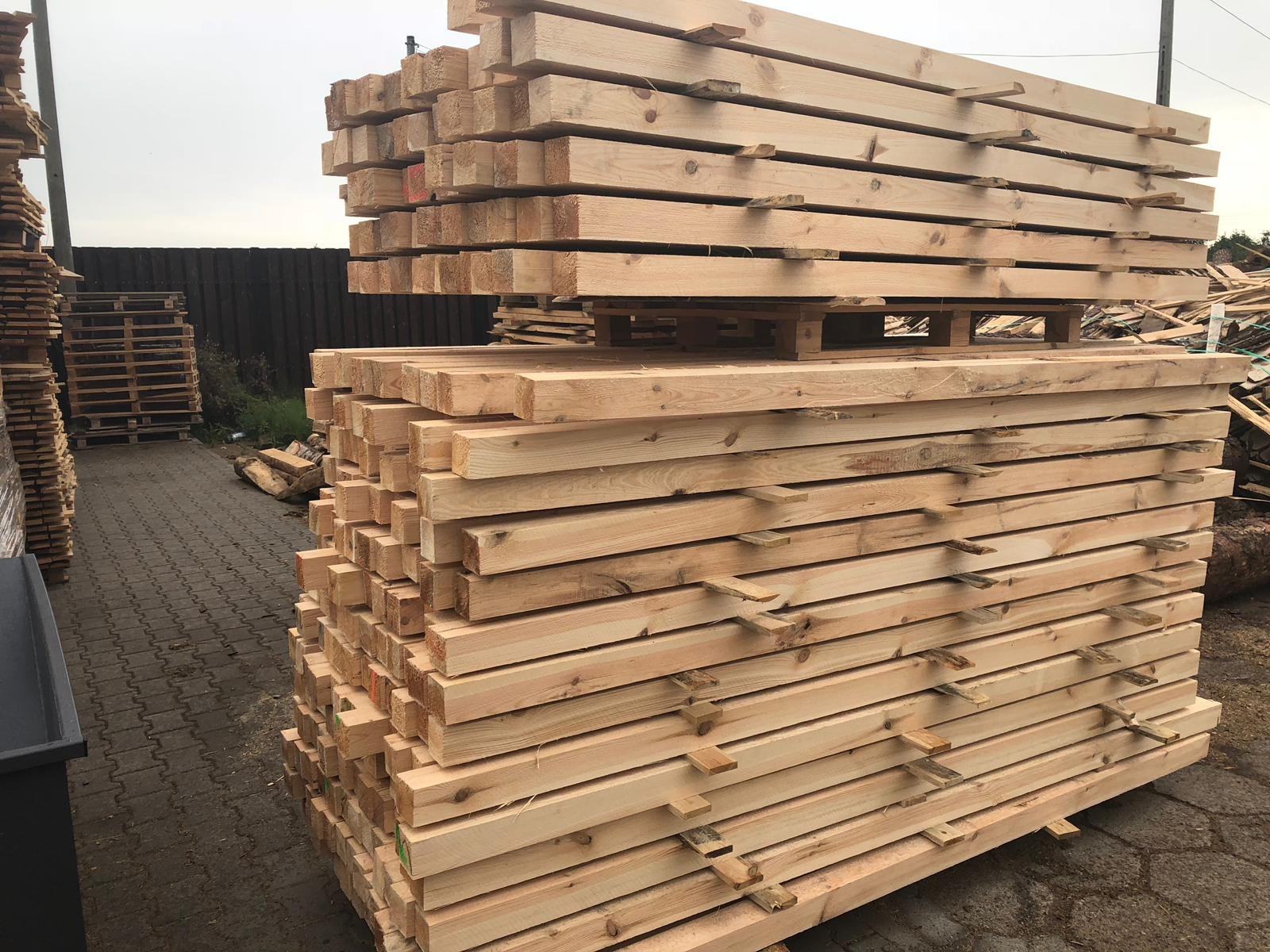 Stos świeżo przetartego drewna budowlanego ułożonego na paletach, widoczne ślady obróbki, w tle ogrodzenie i inne drewniane elementy.