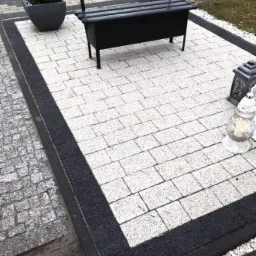 Wyłożony jasną kostką brukową teren z czarną obwódką, na którym stoi czarna ławka i donice z roślinami, w tle widoczny fragment trawnika.