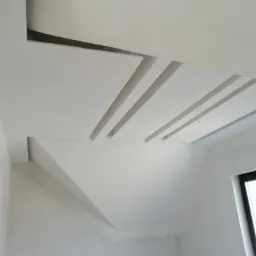 Biały sufit z geometrycznymi wnękami i listwami dekoracyjnymi, widoczne niedoskonałości na ścianach, okno w prawym dolnym rogu.