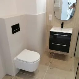 Nowoczesna łazienka z podwieszaną toaletą, czarną armaturą i szafką pod umywalkę oraz lustrem w nieregularnym kształcie, wykończona beżowymi płytkami imitującymi kamień.