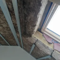 Skośny sufit poddasza z widoczną warstwą izolacji z wełny mineralnej, metalowymi profilami konstrukcyjnymi i oknem dachowym z drewnianą ramą.