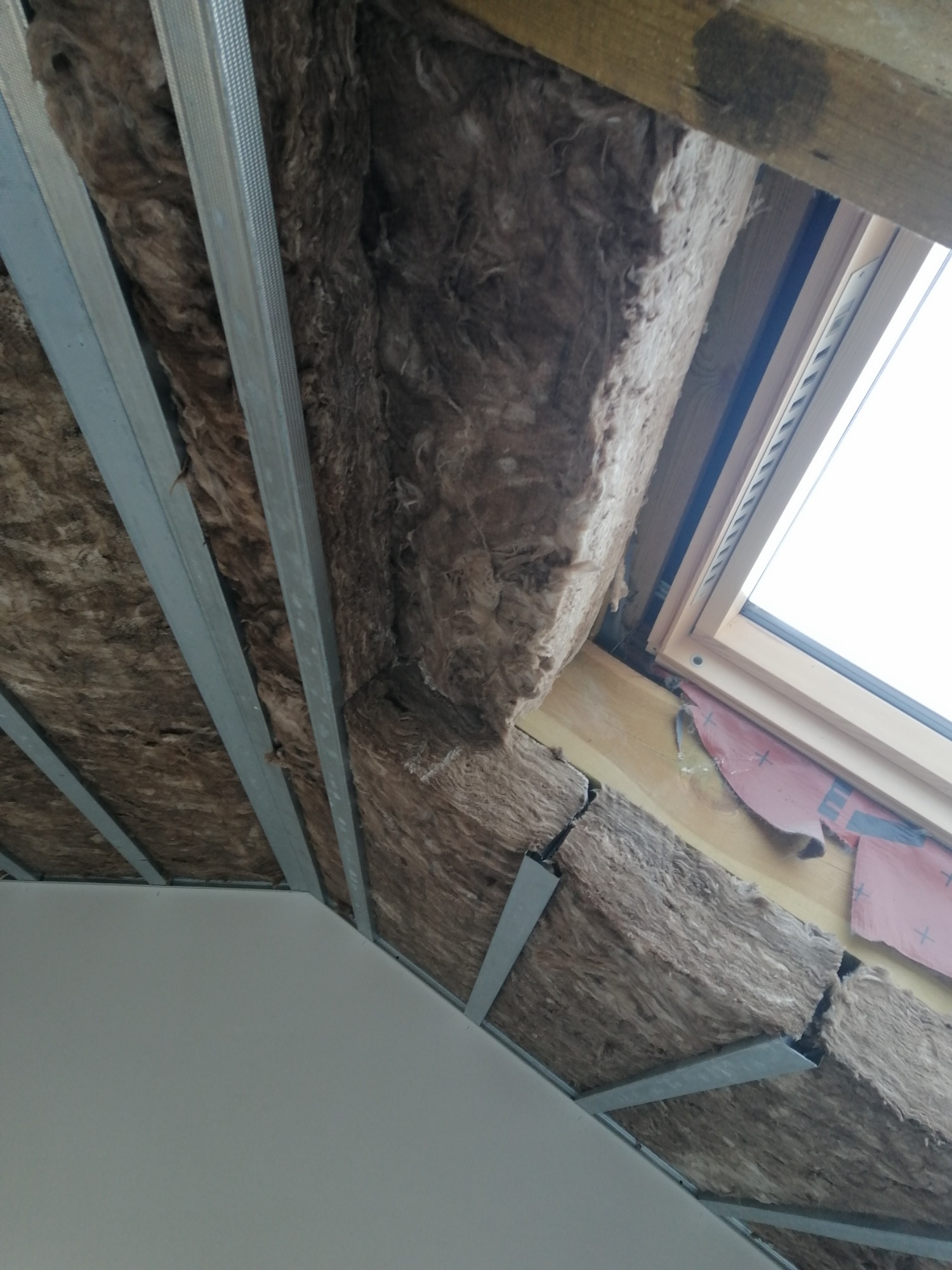 Skośny sufit poddasza z widoczną warstwą izolacji z wełny mineralnej, metalowymi profilami konstrukcyjnymi i oknem dachowym z drewnianą ramą.