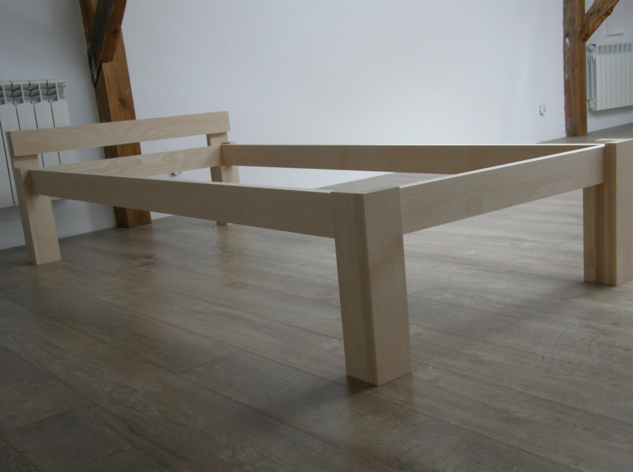 Surowa, drewniana rama łóżka z jasnego drewna na podłodze z paneli, widoczny fragment grzejnika i drewnianej belki stropowej w tle.