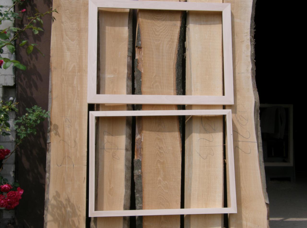 Dwie surowe, jasne ramy okienne ułożone na stosie desek z naturalną korą, z widocznymi numerami ołówkiem i kwitnącym krzewem róży w lewym dolnym rogu.