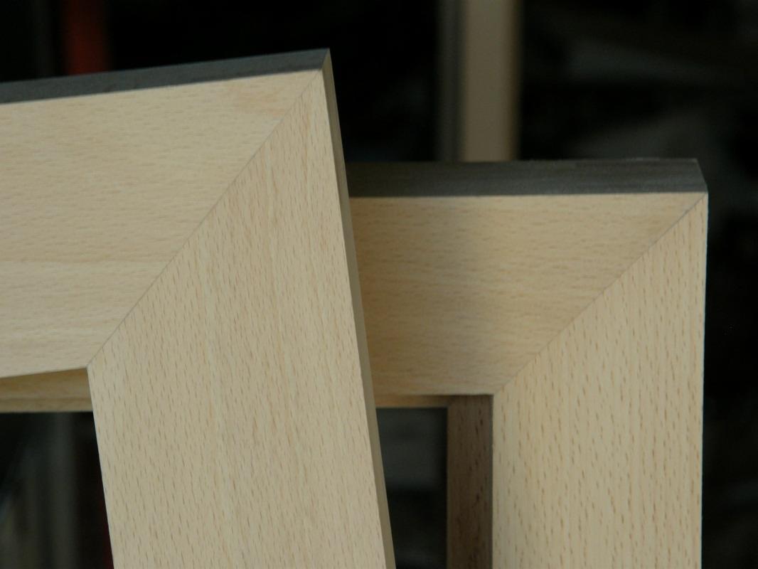 Dwa jasne, drewniane narożniki ram okiennych lub drzwiowych z szarym wykończeniem, widoczne z bliska, na ciemnym tle.