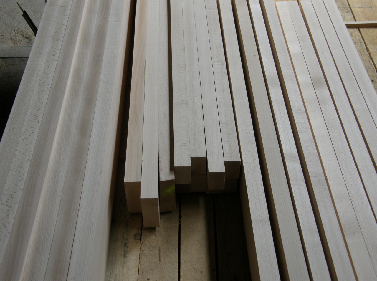 Stos równo ułożonych, surowych kantówek z jasnego drewna na drewnianej podłodze, przygotowanych do dalszej obróbki stolarskiej.