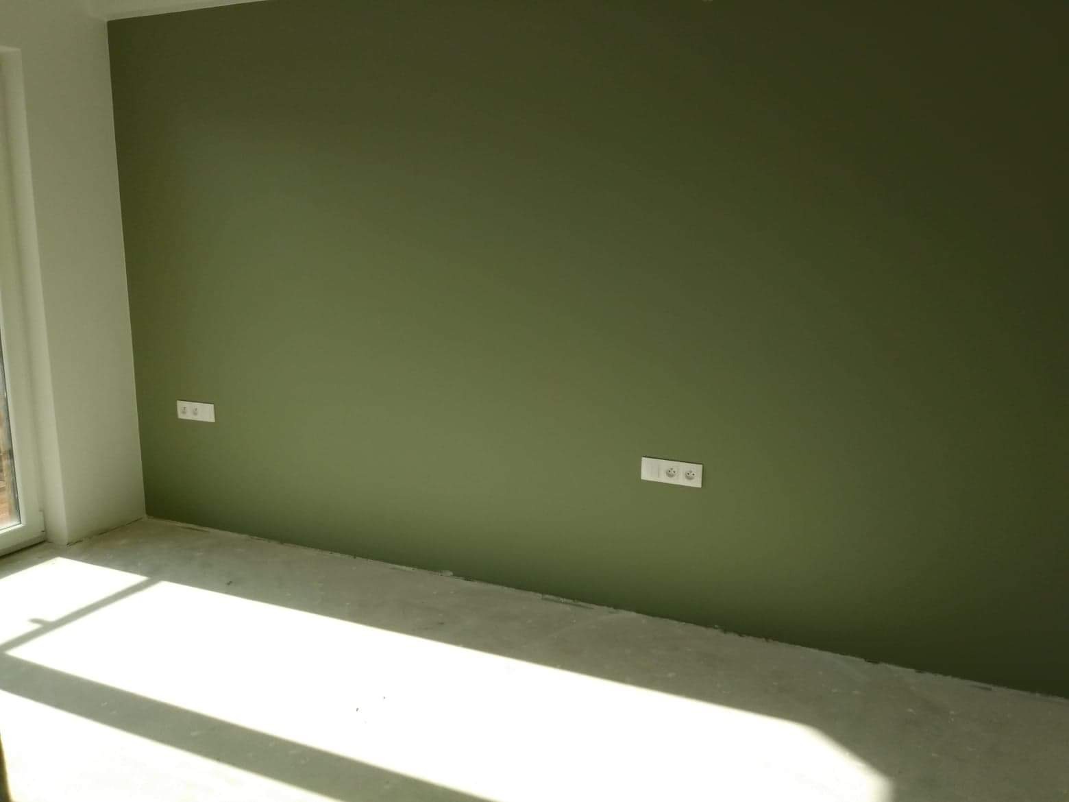Świeżo pomalowana ściana w odcieniu oliwkowej zieleni z zamontowanymi gniazdkami elektrycznymi. Na podłodze widoczne ślady po remoncie i promienie słoneczne wpadające przez okno.