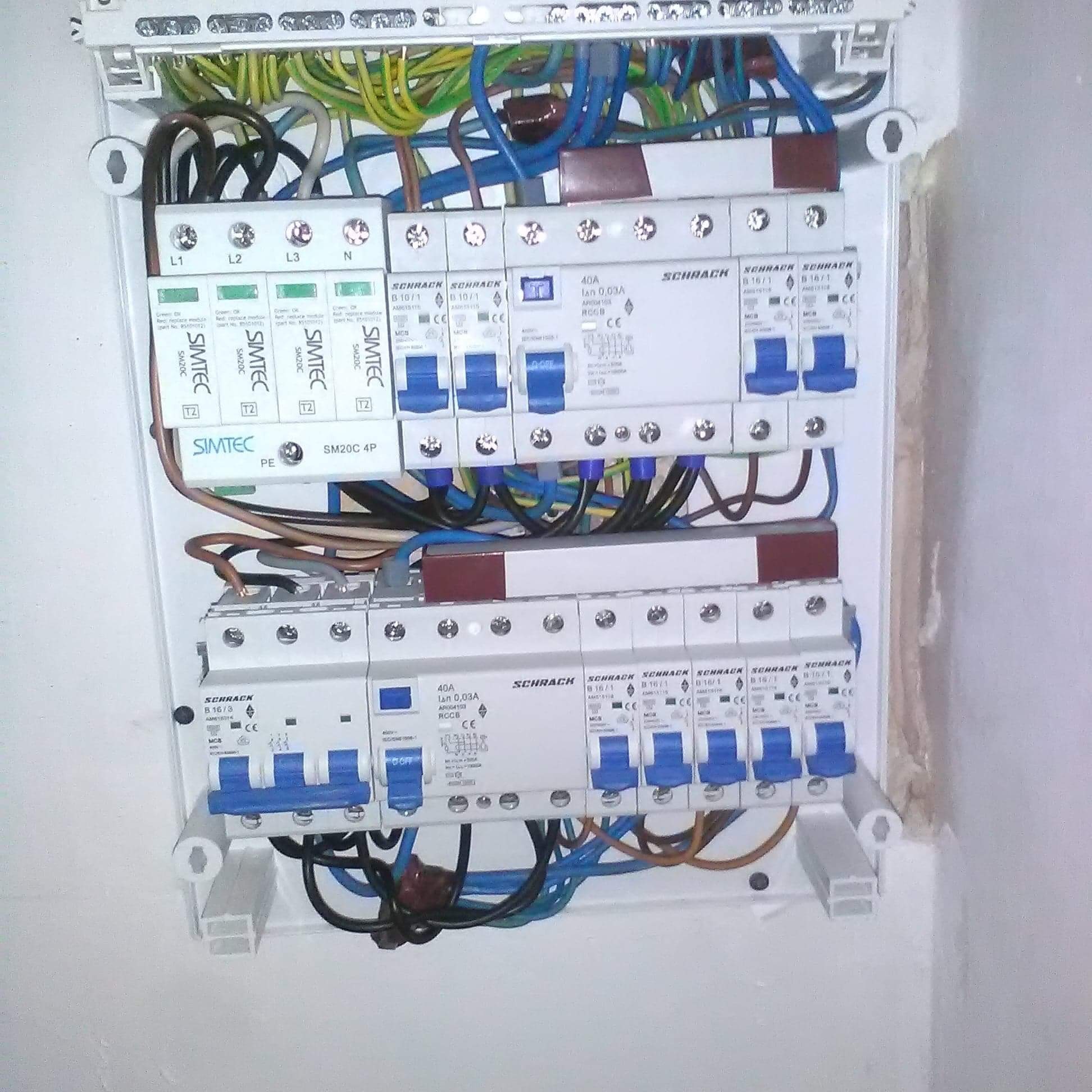 Otwarta skrzynka elektryczna z widocznymi bezpiecznikami, przewodami i elementami zabezpieczeń, zamontowana na białej ścianie.