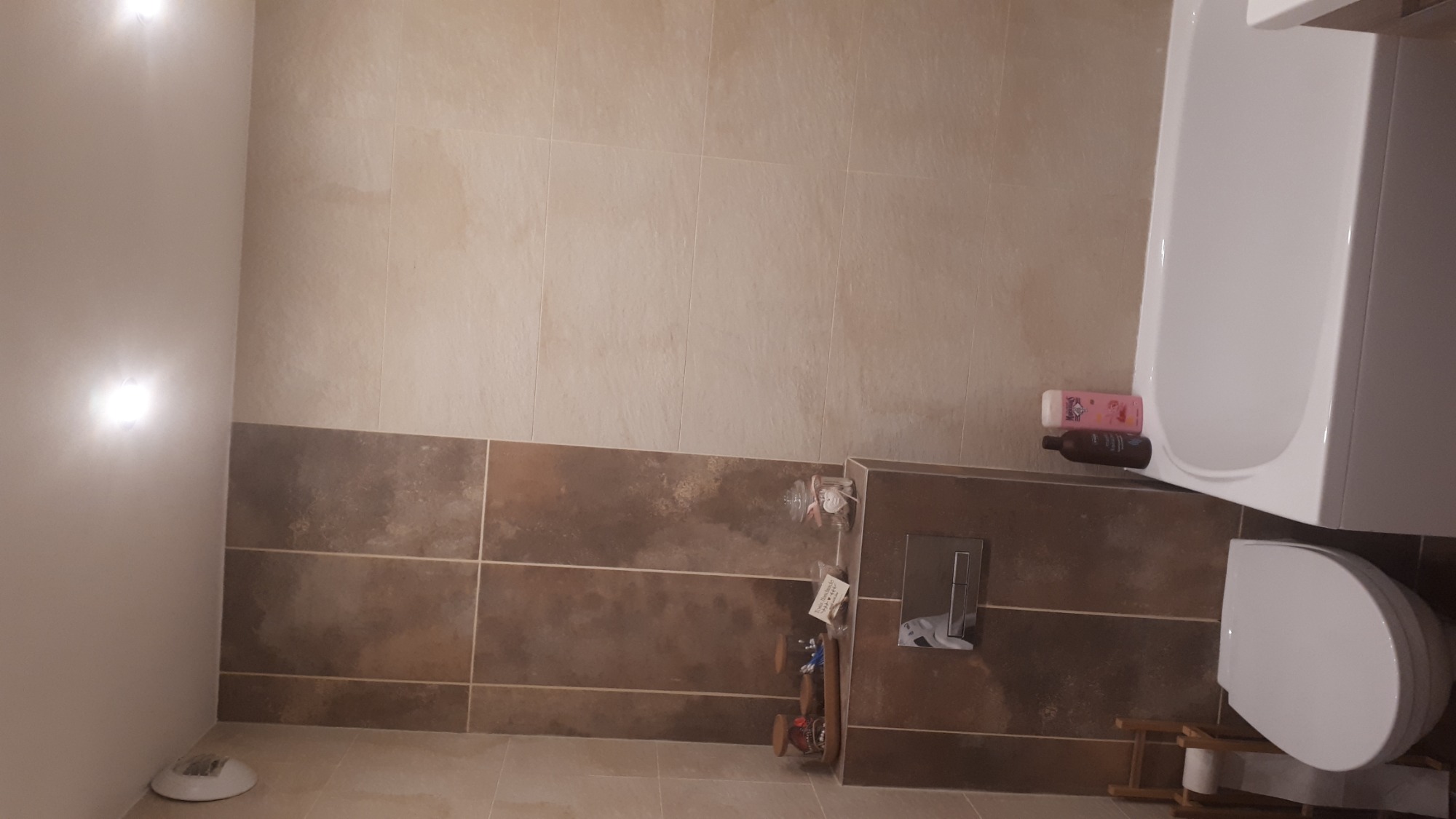 Wykończona łazienka z beżowo-brązowymi płytkami, białą wanną i toaletą; widoczne akcesoria łazienkowe i oświetlenie.