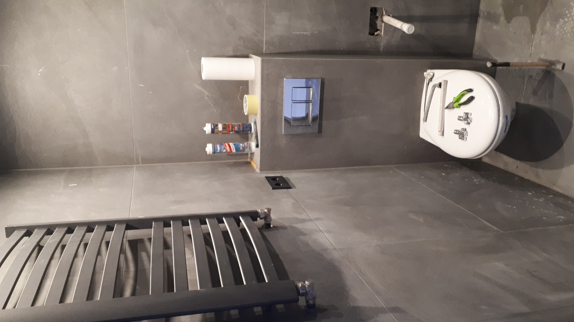 Szare płytki w łazience w trakcie remontu, widoczny grzejnik drabinkowy, podwieszana toaleta z narzędziami na desce, oraz puszki z silikonem i przycisk spłuczki.