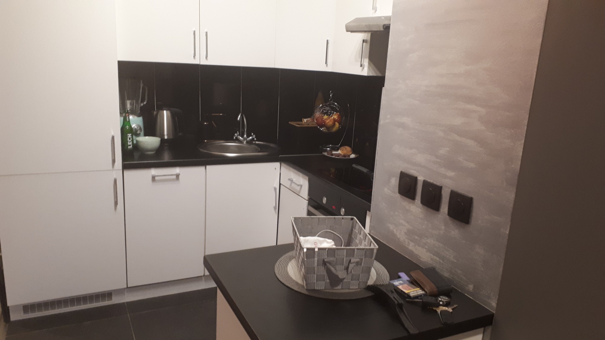 Wykończona kuchnia w Krakowie z białymi szafkami, czarnym blatem i szarą ścianą z efektem dekoracyjnym. Na blacie koszyk z materiału, a na ścianie rząd włączników.