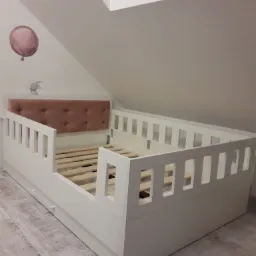 Białe łóżko dziecięce na wymiar z drewnianym stelażem i różowym, pikowanym zagłówkiem, umieszczone na poddaszu ze skośnym sufitem, dekorowane naklejkami balonu i króliczka.