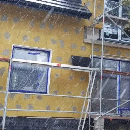 Ściana budynku w trakcie ocieplania, widoczne płyty izolacyjne z szarymi łatami, okna zabezpieczone folią, rusztowanie, padający śnieg.