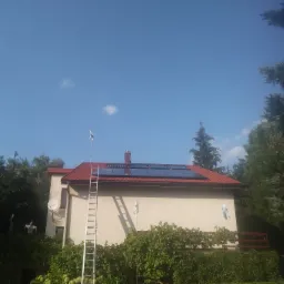 Dom jednorodzinny z panelami słonecznymi na czerwonym dachu, drabina oparta o ścianę, antena satelitarna i mała turbina wiatrowa na dachu, otoczony zielenią w słoneczny dzień.