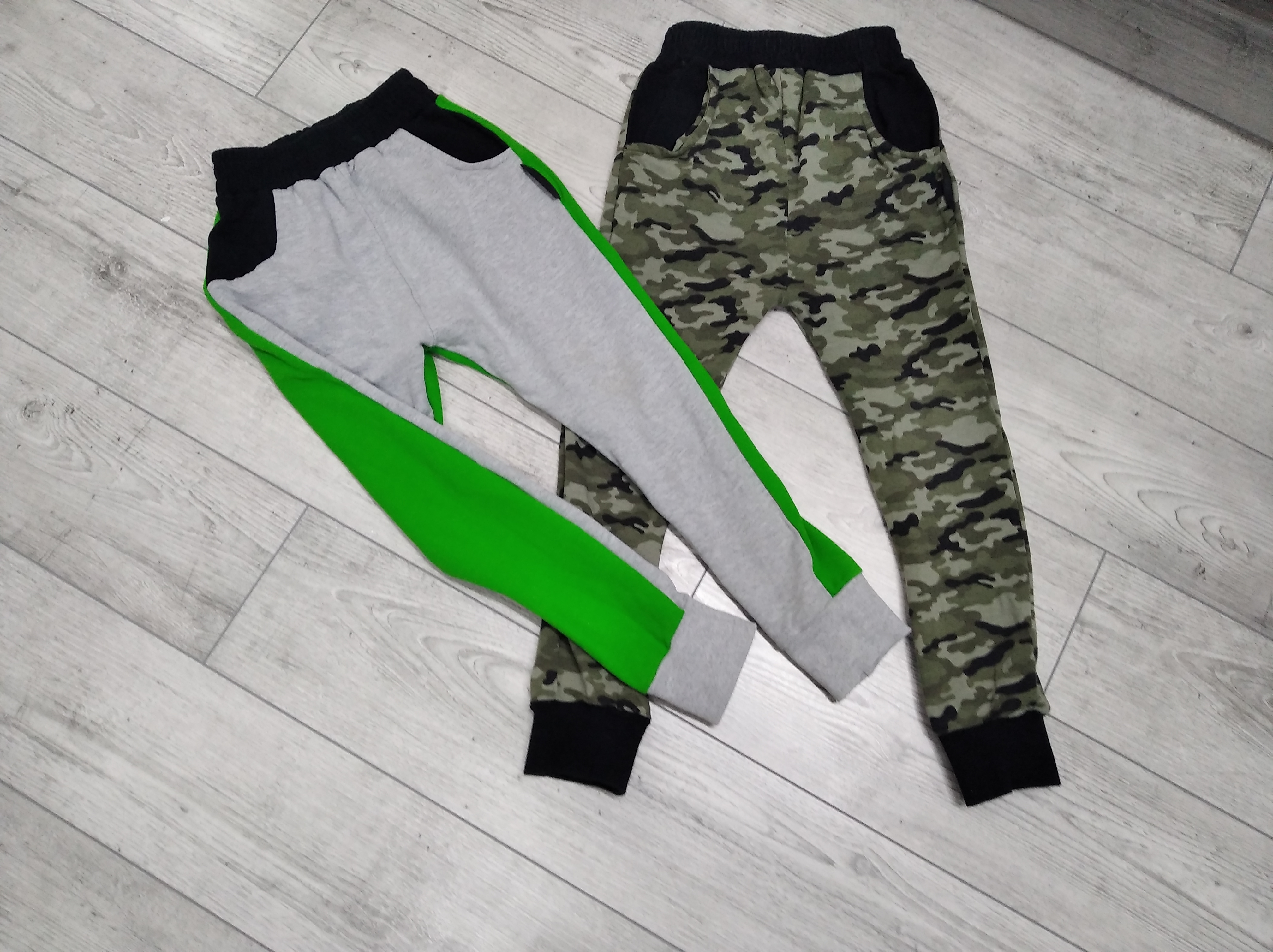 Dwie pary dresowych spodni leżące na podłodze: jedna szara z zielonym paskiem bocznym i czarnym ściągaczem, druga moro z czarnymi kieszeniami i ściągaczem.