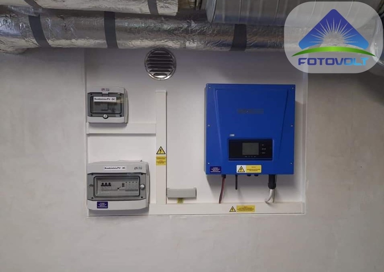 Niebieski falownik fotowoltaiczny zamontowany na ścianie obok dwóch skrzynek zabezpieczeń, widoczne przewody i ostrzeżenia o napięciu, w tle kanały wentylacyjne.