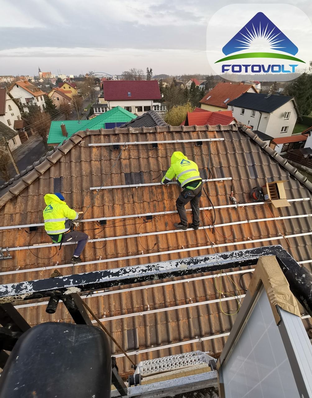 Montaż szyn pod panele słoneczne na dachu z czerwonej dachówki przez dwóch monterów w żółtych kurtkach; widok z góry na tle zabudowy miejskiej i pochmurnego nieba.