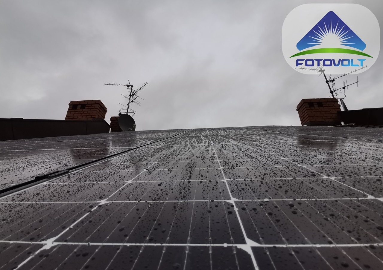 Instalacja paneli słonecznych na dachu pokrytym kroplami deszczu, widoczne kominy i anteny telewizyjne, pochmurne niebo w tle, logo firmy w prawym górnym rogu.