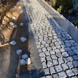 Układanie szarej kostki granitowej na ścieżce ogrodowej, widoczny fragment ułożonej nawierzchni, przygotowane podłoże i rozsypane kostki, obrzeża z betonowych elementów.