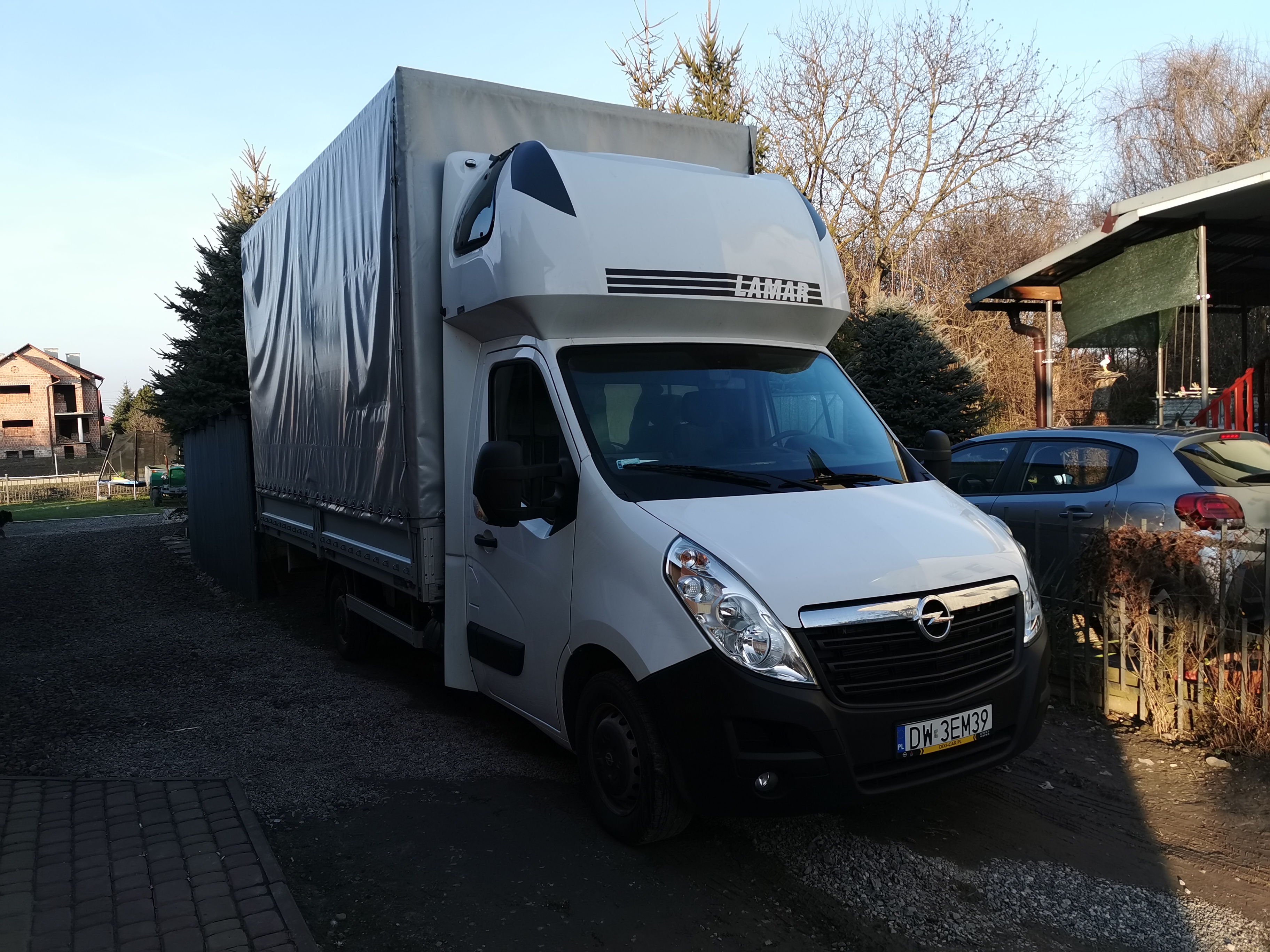 Biały samochód dostawczy Opel Movano z plandeką i nadrukiem 'LAMAR' zaparkowany na żwirowym podjeździe przed domem w słoneczny dzień.